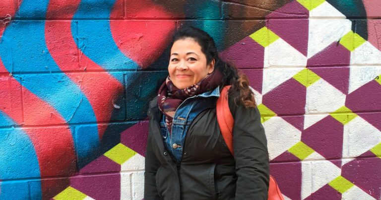 Carol Bilson, dressed in layers, and her backpack, leans against a brightly coloured outside wall.